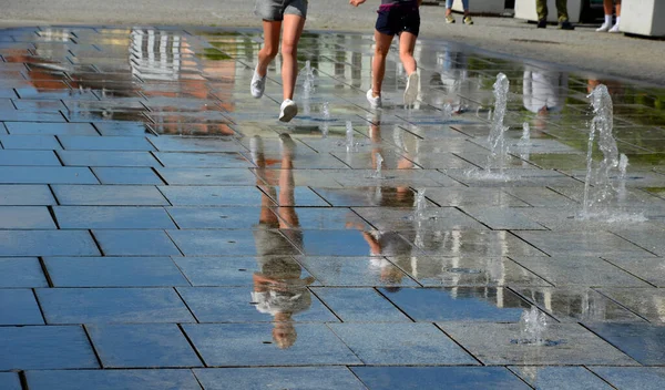 on the square there are spray nozzles in a regular grid which spray upwards according to the program. the girls are fooling around in their shorts and running naked legs through the fountain