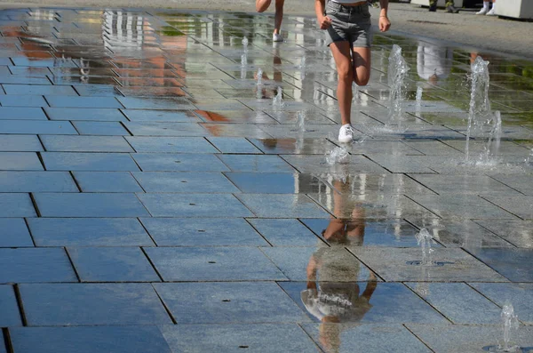 on the square there are spray nozzles in a regular grid which spray upwards according to the program. the girls are fooling around in their shorts and running naked legs through the fountain