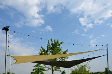 Anaokulundaki oyun parkında, gezinti platformunda güneş ve yağmurun gölgesinde uzanan beyaz tuval. Metalik gri kirişli pergola, kablo, elektrik, ampul,
