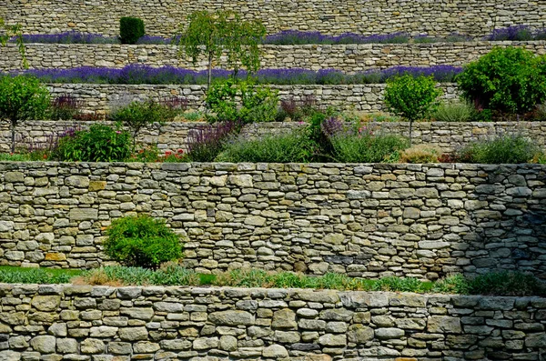 terraces with stairs in a sloping park. stone retaining walls with light stone. blue lavender and pink roses with perennials grow on the edge of the wall. lawns and gravel path, led, footpath, castle