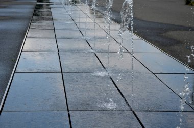 square granite paving stacked in a strip in the sidewalk, silinci, water sprays with alternating spray heights. . water features emerge directly from the cobbled square in the park row. wet tiles on the boardwalk