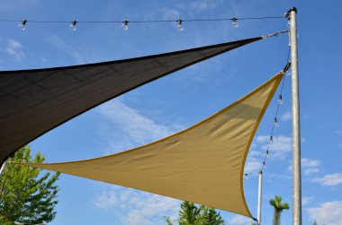 Anaokulundaki oyun parkında, gezinti platformunda güneş ve yağmurun gölgesinde uzanan beyaz tuval. Metalik gri kirişli pergola, kablo, elektrik, ampul,