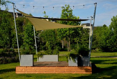 Anaokulundaki oyun parkında, gezinti platformunda güneş ve yağmurun gölgesinde uzanan beyaz tuval. Metalik gri kirişli pergola, kablo, elektrik, ampul,