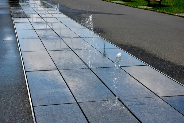 正方形花岗岩铺装在人行道上的一条狭长地带 水喷出交替喷出的高度 是的水的特征直接从公园里的鹅卵石方中显现出来 木板路上的湿瓷砖 — 图库照片