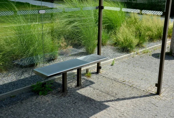 glass bus stop with a metal bench for passengers in the city near the park. the glasses are connected by metal clips. a flower bed with herbs