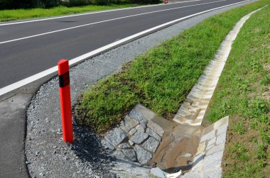 Köprünün altındaki kanalizasyon boruları. Yan yol hendeği geçiyor. Etrafı taşlarla çevrili beton bir delik. Yolun etrafındaki oluklar temiz su, yeni asfalt, kenar ve çakıl dolu.