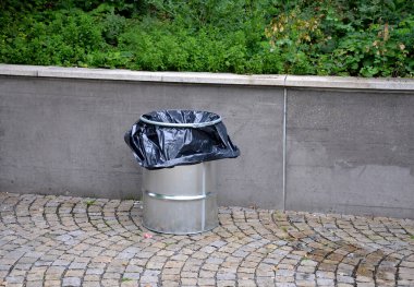 Parktaki beton bir duvarın yanındaki sıralarda. Duvarın üstünde, çiçek tarhında. Metal siyah çerçeveli ahşap park bankı. Çöp torbalı metal varil, konser.