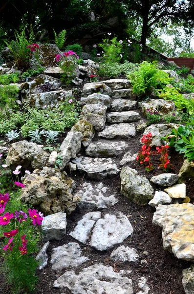 bright limestone staircase on a rock in the garden. steps of rough stones to a hill with flowers. pink red flowers around the path. karst scenery in a historic corner in a romantic park