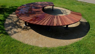 circular bench made of brown-red teak wood. tropical impregnated podia boards for sunbathing visitors in the park. spiral shape, snail shells. trowel base in the lawn, tree, trunk