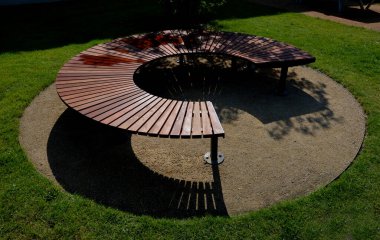 circular bench made of brown-red teak wood. tropical impregnated podia boards for sunbathing visitors in the park. spiral shape, snail shells. trowel base in the lawn, tree, trunk