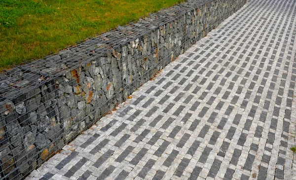 striped white mosaic tile marble. gray stripes sidewalk with walls of gabion baskets around. gray metal garden gate. low fencing for children and dogs. Kindergarten playground