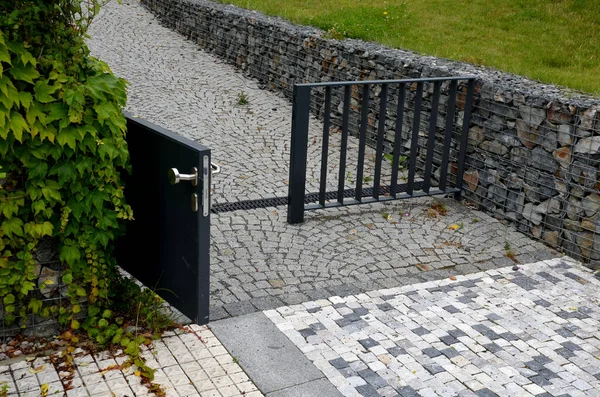 striped white mosaic tile marble. gray stripes sidewalk with walls of gabion baskets around. gray metal garden gate. low fencing for children and dogs. Kindergarten playground