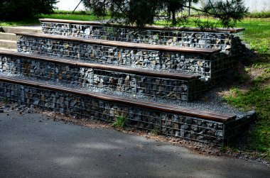 Parktaki tribünde, içinde gri taşlar bulunan Gabion sepetlerinden yapılmış beton bir merdivenin yanında. Koltukların oturma alanları tahtalardan yapılmış basamaklar..