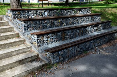 Parktaki tribünde, içinde gri taşlar bulunan Gabion sepetlerinden yapılmış beton bir merdivenin yanında. Koltukların oturma alanları tahtalardan yapılmış basamaklar..