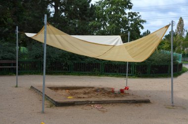 İki tahta sütun arasında bir muşamba var. Çatı, evdeki kum havuzu ya da terasta gölgelendirme görevi görüyor. Ya da ön bahçedeki ve parktaki restoranda. Metal kablolar ve kablolar