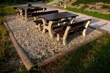 Parktaki gezinti güvertesindeki ahşap banklar. Destek olarak kütüğü olan beton bir bank. Arka planda çok yıllık yataklar ve çimenler var. çakıl dinlenme alanı