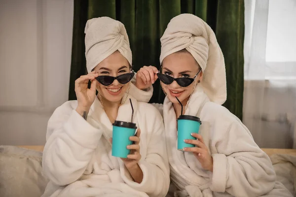 two young women in bathrobes and bathrobe with coffee cups on the sofa in the morning.