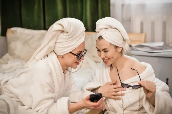 beautiful young women in bathrobe and towel in bathrobes looking at camera while sitting on sofa in spa salon