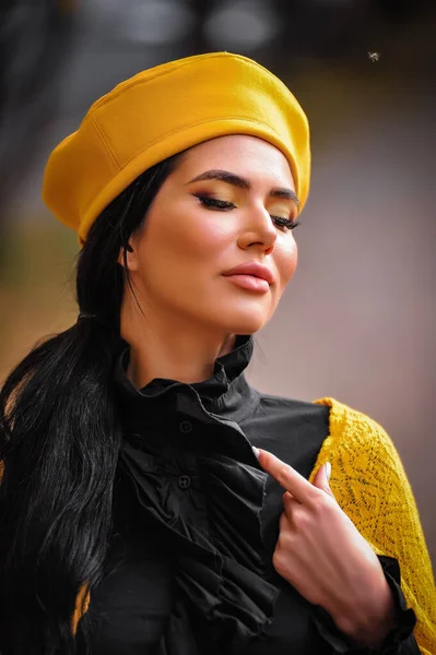 beautiful young woman in a yellow jacket and black hat posing on a background of autumn leaves