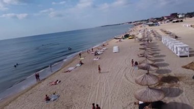 Drone plaj havadan görüntüsü. Kıyı şeridinde bir dron uçuyor: Sazlıklardan yapılmış plaj şemsiyeleri, güneş panzeleri. İnsanlar denizde yüzer, sahilde dinlenir..