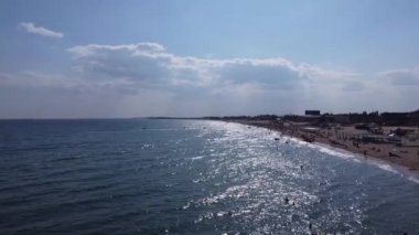 İnsanlar sahilde yüzer. Plaj insanları güneşleniyor, güneşleniyor, Eylül 'de öğleden sonra deniz kenarında dinleniyorlar. Deniz dalgaları ve kum. Orijinal 4K video. Deniz ve kıyı şeridinden insansız hava aracı görüntüsü.