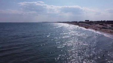 İnsanlar sahilde yüzer. Plaj insanları güneşleniyor, güneşleniyor, Eylül 'de öğleden sonra deniz kenarında dinleniyorlar. Deniz dalgaları ve kum. Orijinal 4K video. Deniz ve kıyı şeridinden insansız hava aracı görüntüsü.