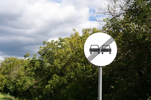 Road sign: End of no overtaking zone. Road Sign canceling of previous limitation. Round road sign with two black cars and black lines against the backdrop of forest and blue sky.