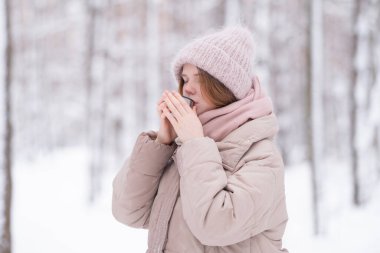 Genç kızıl saçlı bir kadın karla kaplı bir kış ormanında termos bardağından çay içiyor..