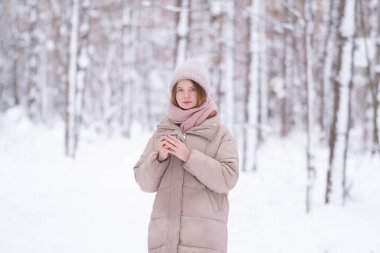 Genç kızıl saçlı bir kadın karla kaplı bir kış ormanında termos bardağından çay içiyor..