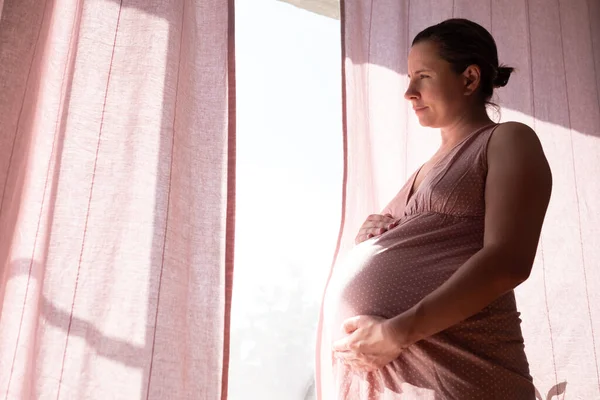 Mulher grávida tocando barriga de pé perto da janela. — Fotografia de Stock