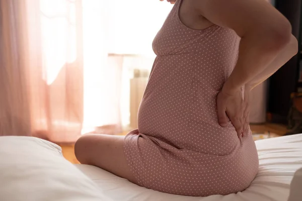 Frau, die zu Hause unter Schmerzen leidet, im Bett sitzt, Bauch und Rücken berührt, werdende Mutter mit Geburtslabo — Stockfoto