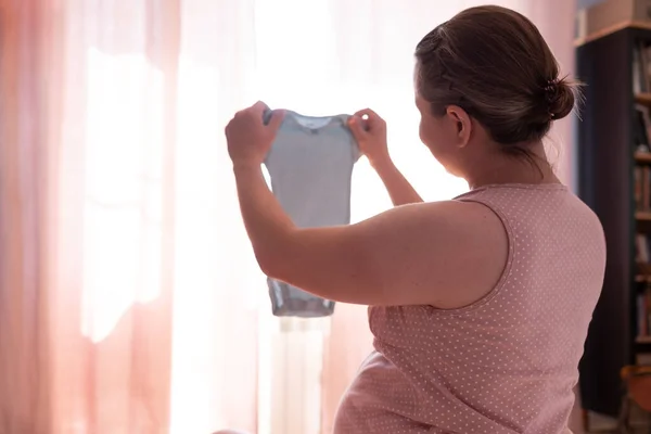 Беременная женщина ищет детскую одежду сидя на кровати подготовки к ребенку — стоковое фото