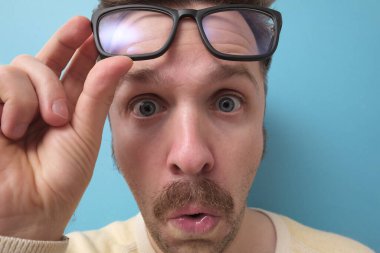 Büyük gözlüklü komik bıyıklı genç adamın şüpheleri var ve mavi arka planda şaşkın bir yüz ifadesi var.