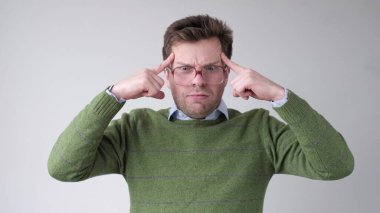 Young funny man in big glasses holds fingers on temples, trying to remember or generate idea. Studio shot clipart