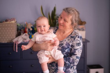 Evde kucağında 7 aylık bir bebek tutan yaşlı bir büyükanne.