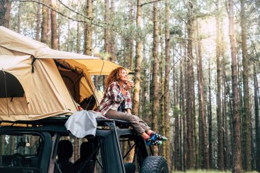 Arabanın çatısında çadırda oturan ve ormanda kahve içen genç bir kadın.. 