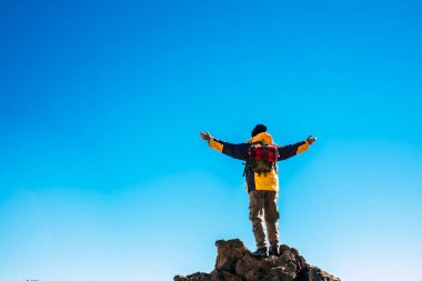 Sırt çantasındaki adamın mavi gökyüzüne uzanmış kollarıyla kayanın üzerinde duruşu..
