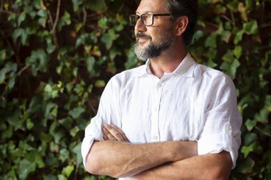 Düşünceli, olgun bir adam, gözlük takıyor ve kollarını yeşil yaprakların önünde kavuşturup arkasını çeviriyor.. 