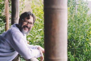 Neşeli, genç, olgun bir adam gülümseyip arka planda yeşil ağaçlar olan balkonda duran açık hava ev portresindeki kameraya bakın. Gözlüklü mutlu insanlar