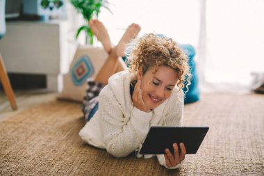 Evdeki halının üzerinde yatan mutlu yetişkin kadın modern tablet ve evdeki internet bağlantısını kullanıyor.. 