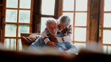 Happy and enjoyed senior caucasian couple smile and hug each other sitting on a chair in cozy home cabin chalet 