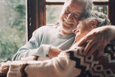 Old senior couple in love hug and embrace with romance together at home with outside view in windows background - happy mature retired people lifestyle enjoying caring each other and smiling
