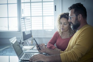 Smart working at home free office lifestyle with adult couple using home and two pc laptop computer on the desktop - modern man and woman online job remote workers bright day light