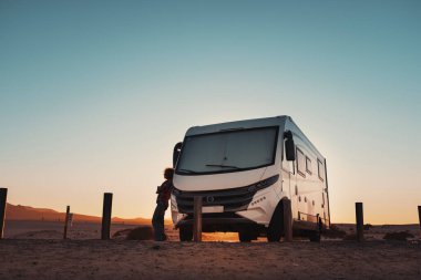 People and camper camping car van freedom concept lifestyle or alternative transport vacation. Big van parked off road and woman resting outside. Colorful sunset and sky with golden mountains in background