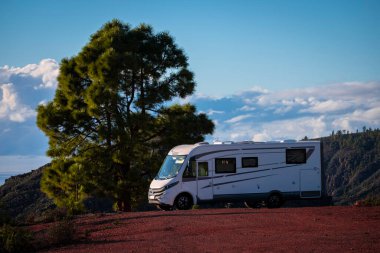 Tatil ve seyahat macera konsepti yaşam tarzı. Büyük bir karavanın karavanı büyük bir ağacın yanında ve arkasında dağlar olan mavi bir gökyüzü.