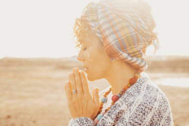 Birleştirilmiş elleri ve kapalı gözleri olan kadın portresi arka planda parlak güneş ışığıyla açık havada dua edip meditasyon yapıyor. Meditasyon ve iç denge kavramı.