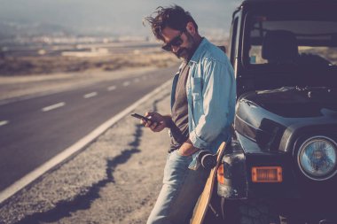 Gezgin adam, arka planda uzun bir yol olan siyah bir off-road aracına karşı duran mobil telefon kullanıyor. Maceraperest erkekler için yolculuk ve seyahat yaşam tarzı. Dijital harita yol gezisi kullanımı