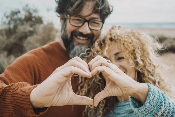 Happy and enjoyed man and woman doing hearth gesture with both hands together. Concept of love and romance with young adult couple enjoying outdoor leisure activity. Beach in background