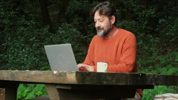 一个满脸胡须的男人在森林里的桌子上工作 一边喝咖啡一边想着工作 在线上网笔记本电脑 — 图库视频影像