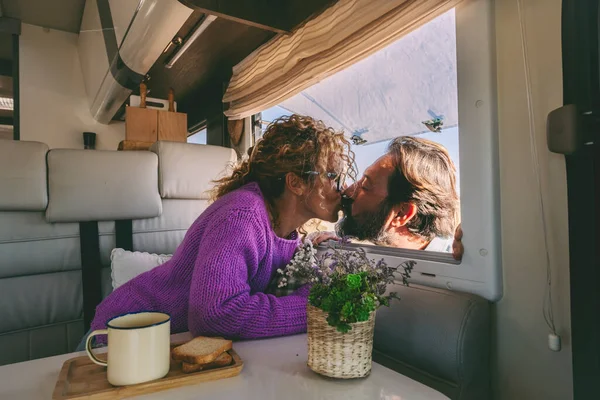 Romantic couple in love kissing during holiday vacation leisure. Woman sitting inside a modern camper kiss her husband outside it. People and vanlife. Relationship in travel summer life.